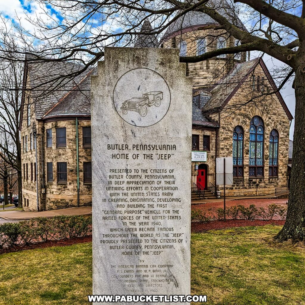 This image features a historical stone monument in Butler, Pennsylvania, proclaiming it as the "Home of the 'Jeep'". The monument has an etched illustration of a classic Jeep at the top and an inscription that reads: "Presented to the citizens of Butler County, Pennsylvania, in deep appreciation of their uniting efforts in cooperation with the United States Army in creating, originating, developing and building the first 'General Purpose' vehicle for the Armed Forces of the United States in the year 1940, which later became famous throughout the world as the 'Jeep'". The text acknowledges the citizens and mentions the American Bantam Car Company, the designers, and engineers involved. In the background, there is a two-story stone building with arched windows, alongside a church with stained glass and a pointed steeple, set against a partly cloudy sky. Bare tree branches frame the image on the left, suggesting it was taken in late autumn or winter.