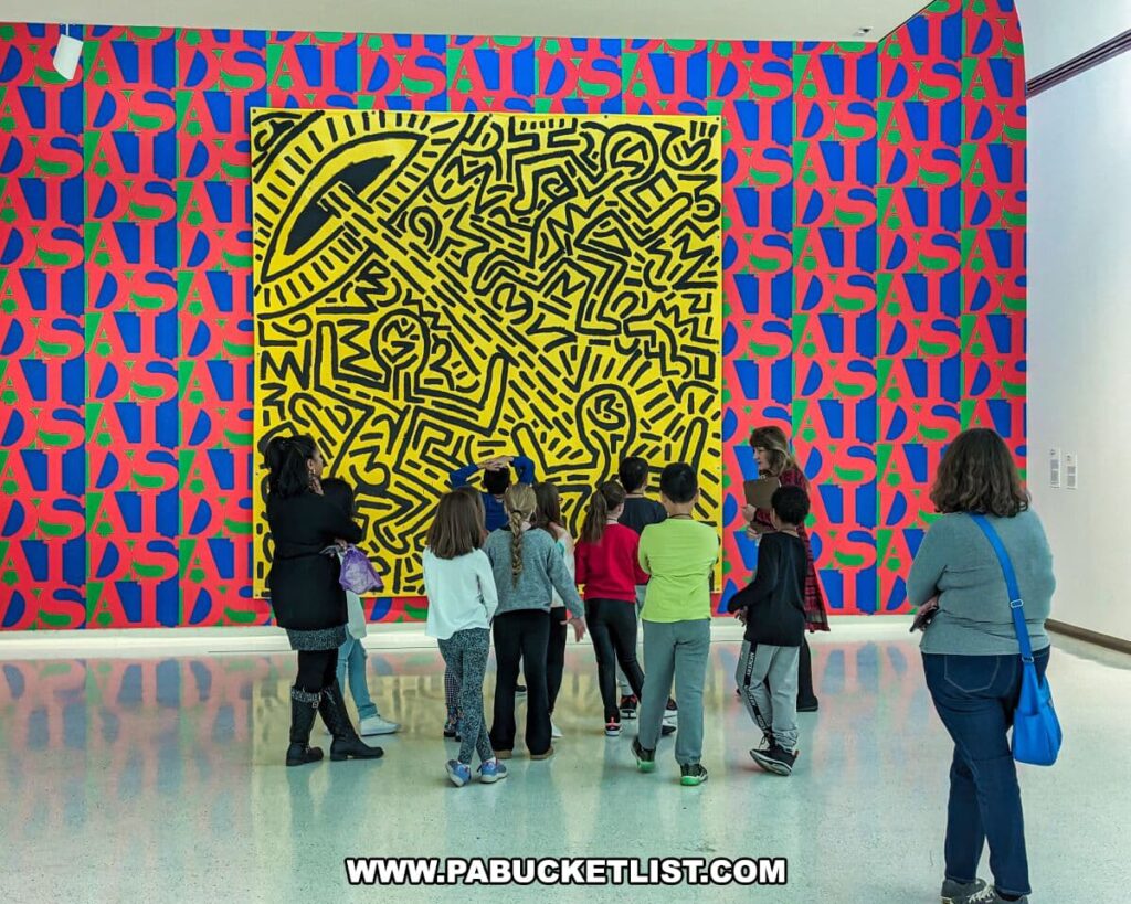 A group of visitors, including children and adults, is engaged with a large, bold artwork at the Carnegie Museums of Art and Natural History in Pittsburgh, PA. The artwork features a dynamic yellow and black labyrinth-like design set against a vibrant background of red, blue, and green with the repeated word 'AIDS.' The viewers are standing, some pointing, as they examine and discuss the piece. The bright artwork contrasts with the museum's sleek, white interior space.