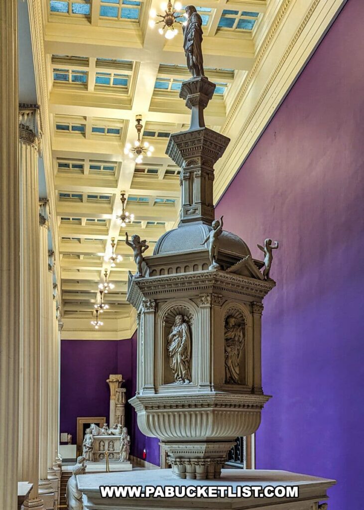 A photograph from the Hall of Architecture at the Carnegie Museums of Art and Natural History in Pittsburgh, PA, showing an elaborate plaster cast of a baptismal font. The font is adorned with sculpted figures and classical motifs, capped with a statue of John the Baptist. It is set against the backdrop of a coffered ceiling and classical columns, with a purple wall accentuating the intricate details of the cast. The exhibit highlights the museum's collection of architectural replicas and provides visitors with an up-close view of historical artistry.