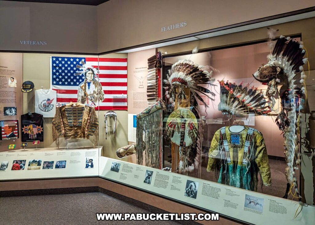 An exhibit dedicated to Native American veterans at the Carnegie Museums of Art and Natural History in Pittsburgh, PA, displaying traditional headdresses and attire. The items are showcased against a backdrop of the American flag with an indigenous person's image superimposed on it, symbolizing the intersection of Native American heritage and military service. Various memorabilia, such as beaded vests and shirts, along with information panels, provide a narrative of the contributions and sacrifices of Native American veterans.