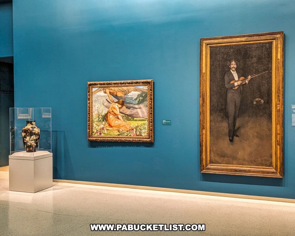 An art exhibit at the Carnegie Museums of Art and Natural History in Pittsburgh, PA, features a serene gallery space with teal blue walls. On the left is a classical painting of a reclining nude in a pastoral setting, framed in gold. To its right hangs a portrait of a man playing the violin, also framed in an ornate gold frame. Between these works is a sculptural vase with a floral arrangement, displayed in a glass case on a pedestal. This curated arrangement of artworks offers a visual harmony and invites contemplation from viewers.