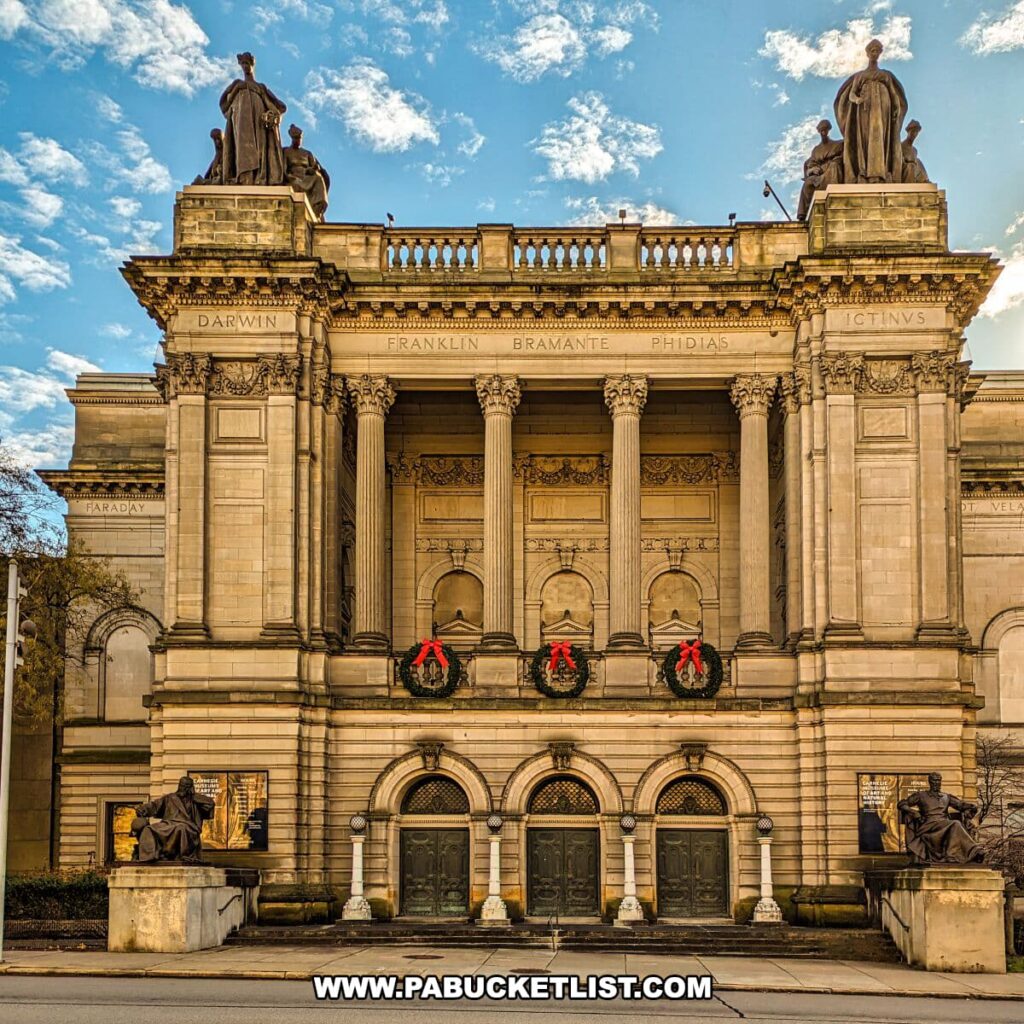 The classical facade of the Carnegie Museums of Art and Natural History in Pittsburgh, PA, adorned with holiday wreaths on its grand entrance. Statues of prominent historical figures, including Darwin and Franklin, are perched atop the building, while the names of other influential figures such as Bramante and Phidias are inscribed on the entablature. Sculptures flank the steps leading to the ornate bronze doors, and the architectural grandeur of the museum is highlighted by the soft glow of the afternoon sun.