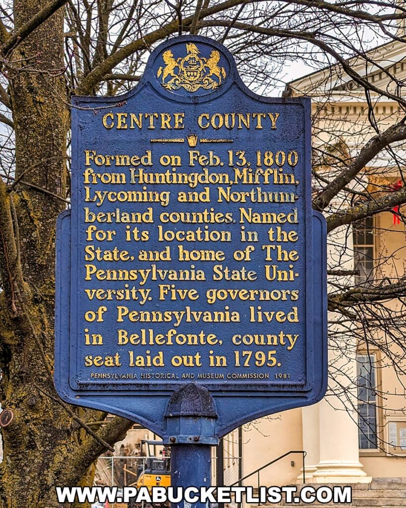 A historical marker in Centre County, Pennsylvania. The sign is blue with raised gold lettering and a decorative crest at the top. It reads: "CENTRE COUNTY. Formed on Feb. 13, 1800, from Huntingdon, Mifflin, Lycoming and Northumberland counties. Named for its location in the State, and home of The Pennsylvania State University. Five governors of Pennsylvania lived in Bellefonte, county seat laid out in 1795. PENNSYLVANIA HISTORICAL AND MUSEUM COMMISSION 1981." Behind the sign, a leafless tree and the corner of a classical-style building with columns can be seen.