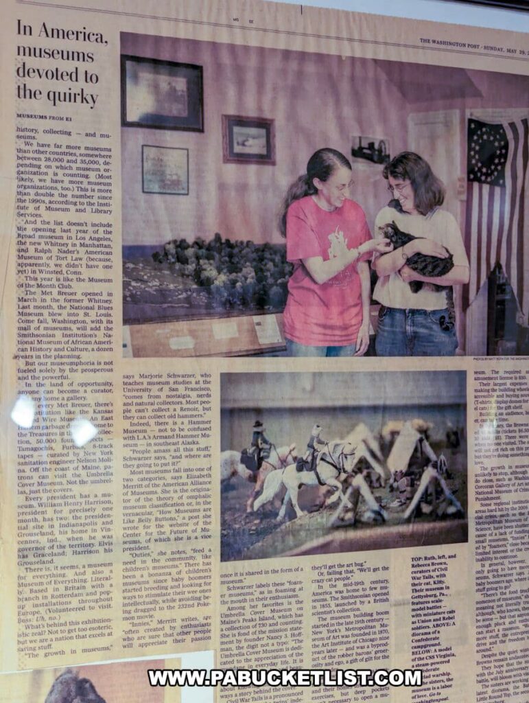 A photo of a newspaper article from 'The Washington Post' displayed at the Civil War Tails Diorama Museum in Gettysburg, PA. The article, titled 'In America, museums devoted to the quirky,' features an image of two women, presumably the museum's founders, holding a cat and smiling. Below them is an image of a diorama depicting a cavalry charge with figures replaced by cats. The text of the article, discussing various unique museums, is not fully legible in the photo. This exhibit highlights the museum's recognition for its distinctive approach to historical dioramas.