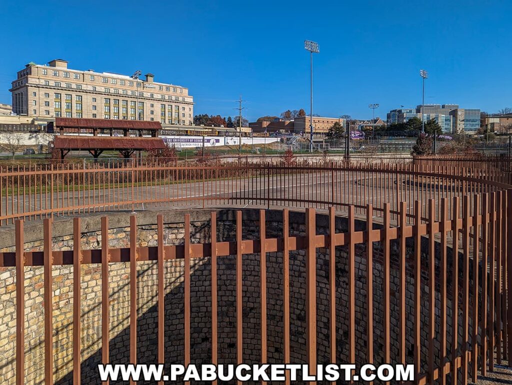 A clear day view of the Scranton Iron Furnaces Historic Site in Scranton, PA, featuring a rust-colored metal fence in the foreground with a stone foundation, a gazebo-style structure in the middle ground, and the historic DL&W Train Station, now a hotel, in the background against a blue sky. Sports fields and modern buildings are visible further back.