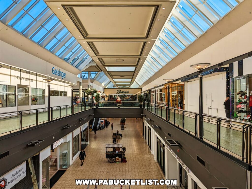 Interior of The Marketplace at Steamtown in downtown Scranton, Pennsylvania, featuring a spacious, brightly-lit mall with a glass ceiling skylight. Storefronts line the multi-level shopping area, including a 'Geisinger' therapy services clinic. Shoppers are visible on both levels, and kiosks dot the ground floor. The atmosphere is calm with modern décor and clean, open spaces.