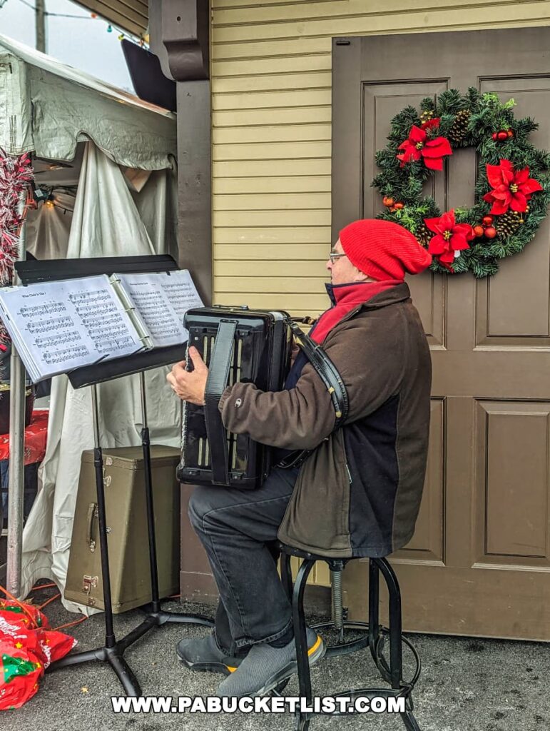A musician in a red beanie and brown coat plays the accordion while seated on a stool outside a train station. He is positioned in front of a music stand with sheets of music, likely playing Christmas carols, evidenced by the festive wreath with red ribbons on the door behind him. The setting appears to be a holiday event, given the seasonal decorations and the musician's attire. The atmosphere seems cheerful and welcoming, inviting passersby to enjoy the live music during the steam train excursion at the Everett Railroad in Hollidaysburg, Pennsylvania.