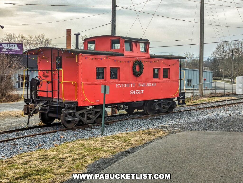 A vibrant red caboose with yellow trim and the words "EVERETT RAILROAD 91517" sits on the tracks in Hollidaysburg, Pennsylvania. It features a black wreath with a bow on its side, adding a festive touch. The caboose is equipped with railings, ladders, and a raised lookout area, characteristic of traditional caboose design. In the background, there's a sign for an outlet store, and various buildings, power lines, and bare trees indicative of a small industrial area. The sky is overcast, and the ground appears dry, suggesting a cool, cloudy day.