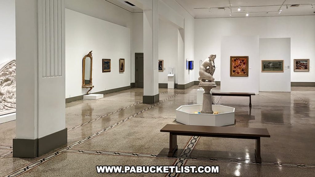 A spacious art gallery within the Everhart Museum in Scranton, Pennsylvania. Central to the room stands a classical sculpture on a pedestal, surrounded by various framed artworks adorning the walls. On the right, an elegant mirror adds depth to the space. The gallery is designed with a terrazzo floor and soft lighting, creating a serene environment for art appreciation. Benches are strategically placed for visitors to sit and engage with the exhibits.