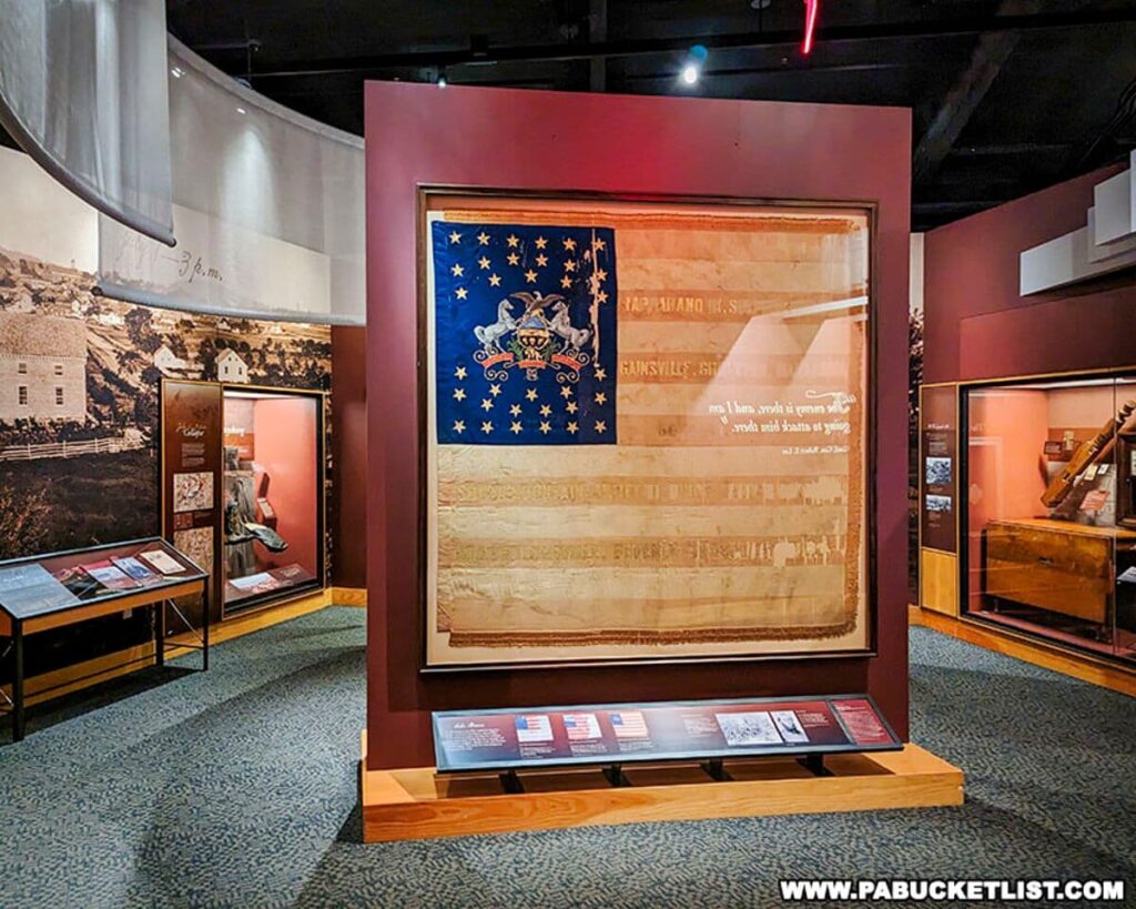 Exhibit at the Gettysburg National Military Park Visitor Center featuring a large, framed battle flag with a circle of stars on a blue canton and faded stripes. The flag is displayed prominently within a protective case against a maroon panel. Around the flag, the exhibit includes descriptive text and smaller displays with additional artifacts. Historical photographs and information panels can be seen in the surrounding area, providing a comprehensive view of the Civil War era.