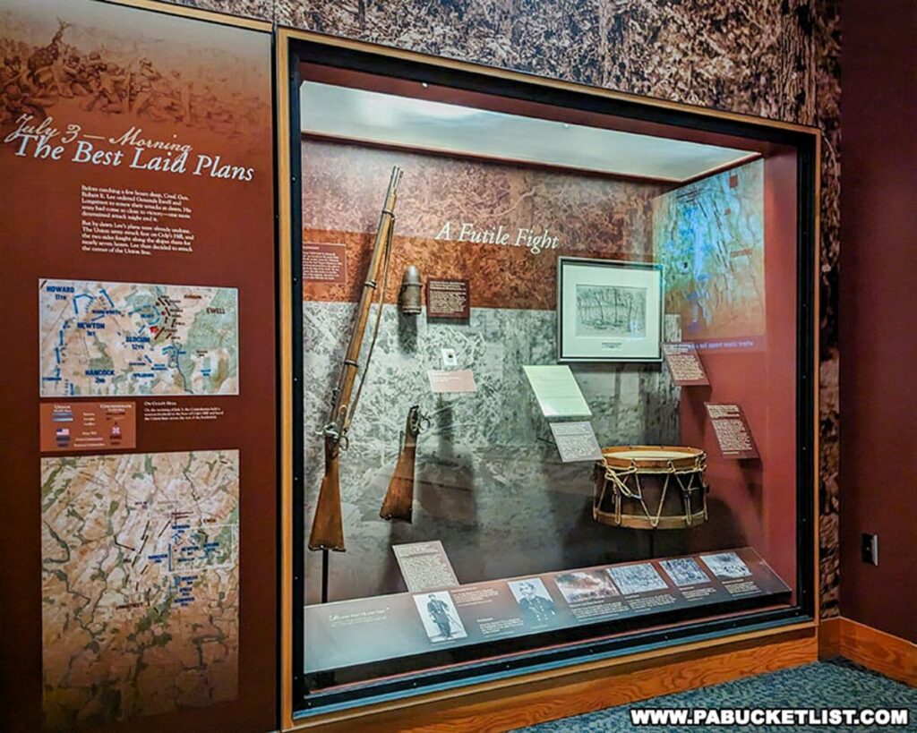 An exhibit display at the Gettysburg National Military Park Visitor Center titled 'July 3 – Morning The Best Laid Plans' and 'A Futile Fight'. It features maps, a rifle, a drum, and informational panels detailing troop movements and strategic plans of the Battle of Gettysburg. The exhibit provides historical insights into the battle's unfolding and key figures involved, presented on a backdrop that replicates the textures and colors of the battlefield.