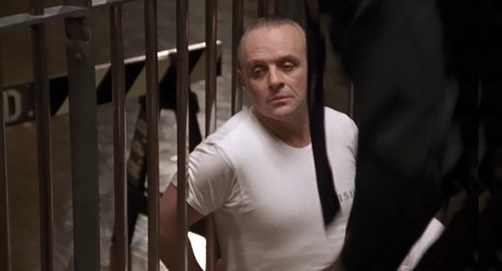 A still from the film "The Silence of the Lambs" showing the character Dr. Hannibal Lecter. He is portrayed as sitting in a prison cell, gazing outward with a contemplative expression. Lecter is wearing a plain white T-shirt and a sling on his arm. The setting features the recognizable metal bars of a prison, emphasizing confinement. This scene was filmed at the Soldiers and Sailors Memorial Hall and Museum in Pittsburgh, PA.