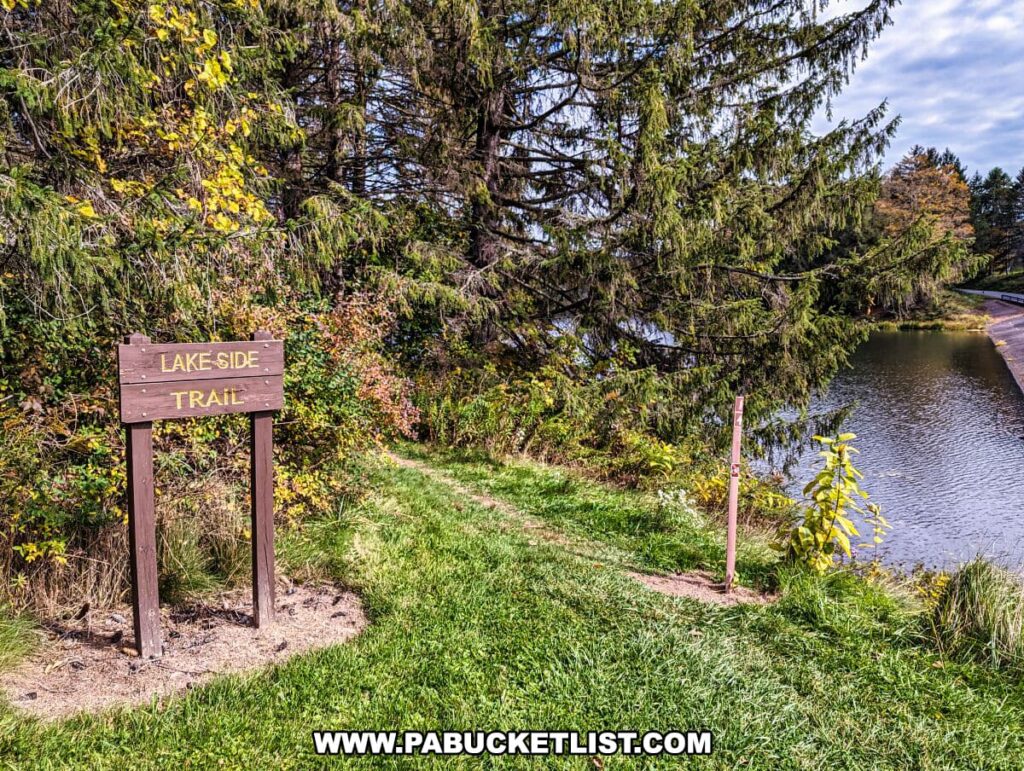 The photo captures the entrance to the "Lakeside Trail" at Hills Creek State Park in Tioga County, Pennsylvania. A wooden sign with the trail name is prominently displayed in the foreground, marking the start of a path that hugs the edge of a calm body of water. The trail seems to lead into a wooded area, with a variety of trees that are beginning to show autumn colors. The grass is lush and green, indicating it's either spring or early fall. The scene is peaceful, with no visible people, suggesting a quiet spot for a walk or contemplation by the lake.