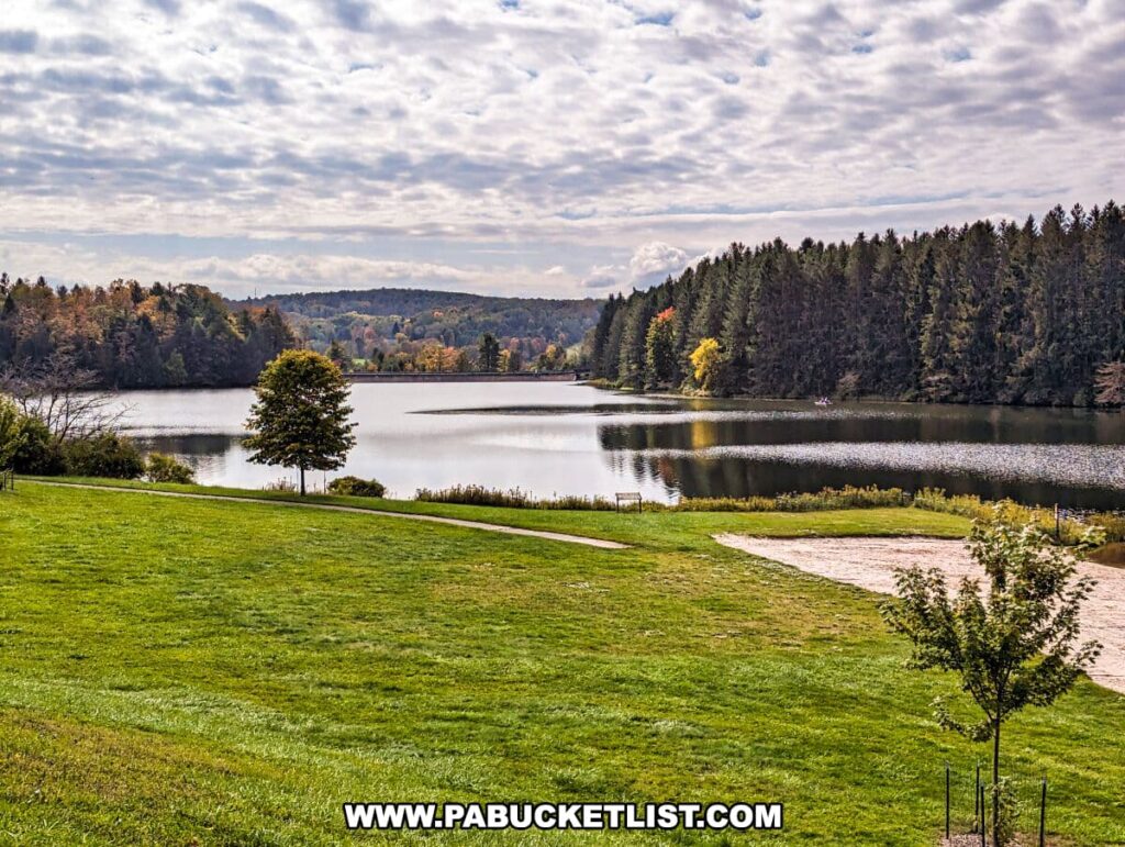 A scenic view of Hills Creek State Park in Tioga County, Pennsylvania. The photo showcases a tranquil lake surrounded by lush forests with the trees beginning to show autumnal colors. A well-maintained green lawn stretches towards a sandy beach at the water's edge. The sky above is partly cloudy, suggesting early fall weather, and the peaceful setting is devoid of people, emphasizing the natural beauty of the area.