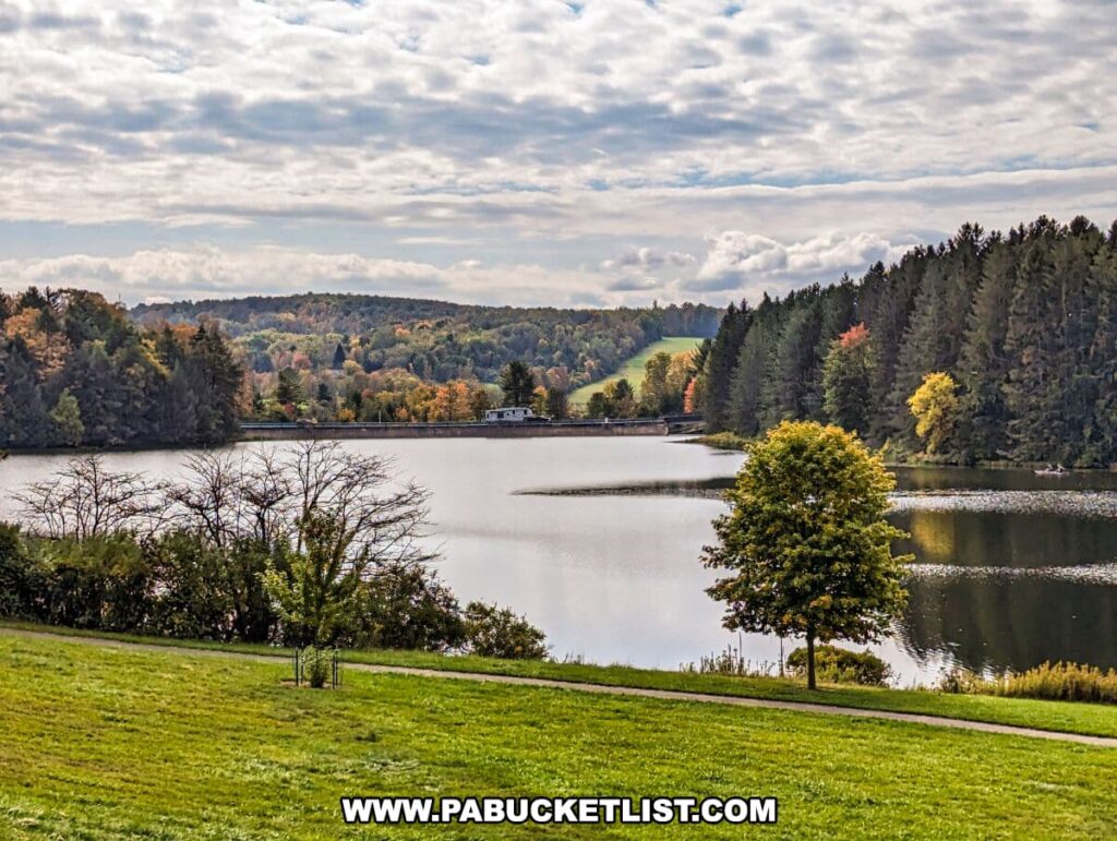 A serene landscape at Hills Creek State Park in Tioga County, Pennsylvania. The vantage point is from a hilltop overlooking a calm lake that mirrors the partly cloudy sky above. Various shades of early fall colors dot the dense forest surrounding the water. In the distance, a dam is visible with a camper parked near it, and a boat can be seen on the lake, adding a touch of recreational activity to the scene. Two prominent trees in the foreground, one bereft of leaves and the other in full green, frame the picturesque view. The overall setting conveys a tranquil autumn day perfect for outdoor activities and enjoying nature.