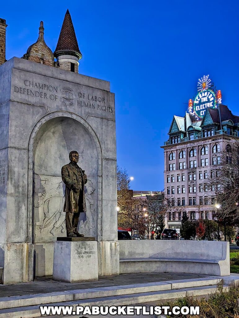 A monument of John Mitchell, labeled 'CHAMPION OF LABOR, DEFENDER OF HUMAN RIGHTS' stands in downtown Scranton, Pennsylvania. The bronze statue is set within a large stone arch, with the name 'JOHN MITCHELL 1870-1919' at the base. In the background, the distinctive Electric City sign illuminates atop a historic building against the evening sky.