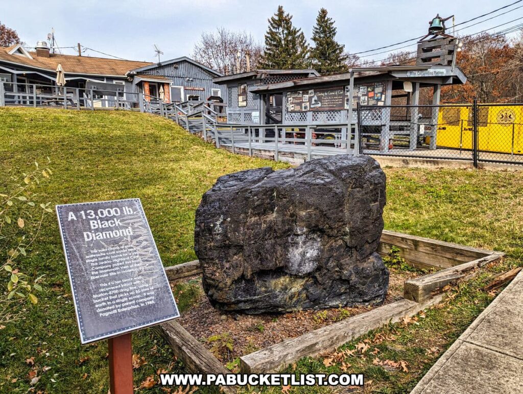 A large 13,000-pound chunk of coal on display at the Lackawanna Coal Mine Tour in Scranton, PA, with an informational sign in front and the coal mine tour's gray wooden building and fencing in the background.