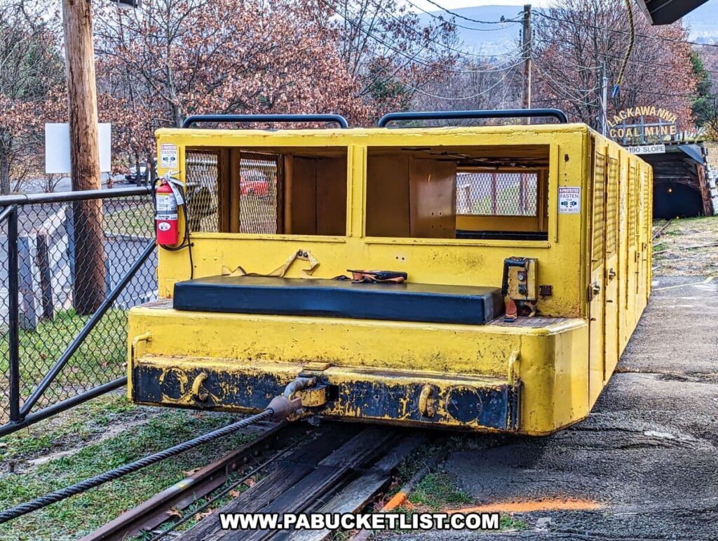 Bright yellow mine car on tracks at the Lackawanna Coal Mine Tour in Scranton, PA. The car, which appears to be an old, rugged vehicle used for transporting miners underground, has a metal grated window and a 'Fasten Seat Belt' sign on its side. It's parked on rails with a forested backdrop and the entrance to the mine visible in the distance, complete with the coal mine's sign.