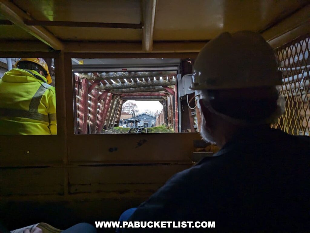 View from inside a mine car looking out onto the Lackawanna Coal Mine Tour in Scranton, PA. Two visitors wearing hard hats are seen from behind, one in a high-visibility jacket and the other in a dark coat. They are gazing out through a window with a metal grid, with the mine's red metal tunnel structure leading out to the daylight where buildings and bare trees are visible.