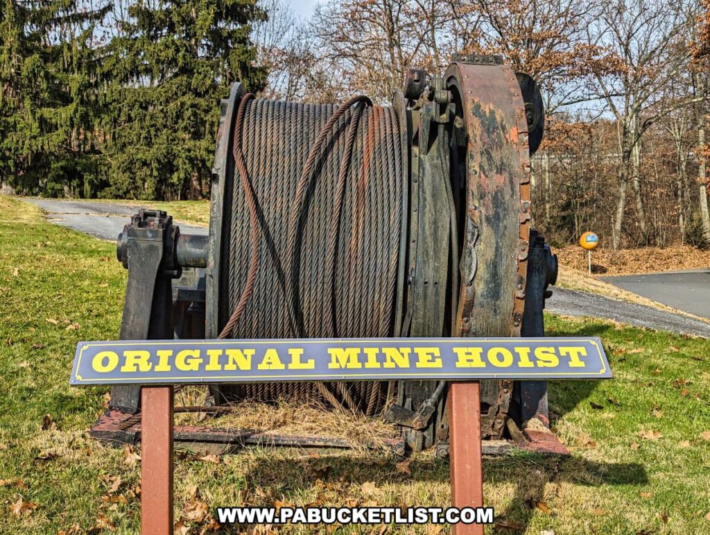 Outdoor display of the 'ORIGINAL MINE HOIST' at the Lackawanna Coal Mine Tour in Scranton, PA. The hoist comprises a large, rusted metal drum around which an array of thick, coiled cables are wound. The machinery sits on a grassy area with trees and a clear sky in the background, showcasing a piece of the historical equipment used in mining operations.