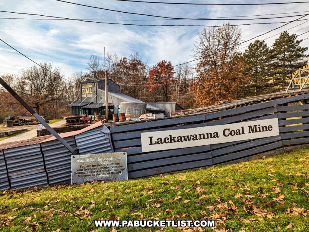 Exterior view of the Lackawanna Coal Mine Tour entrance in Scranton, PA, featuring a large sign with 'Lackawanna Coal Mine' written on it. In the background, there's a gray building with 'COAL MINE TOUR' on its facade, a rusty mining cart on display, and the start of the mine shaft with its yellow structure. The foreground shows a thank-you plaque for visitors set on a corrugated metal fence, amidst a lush setting with autumn-colored trees under a partly cloudy sky.