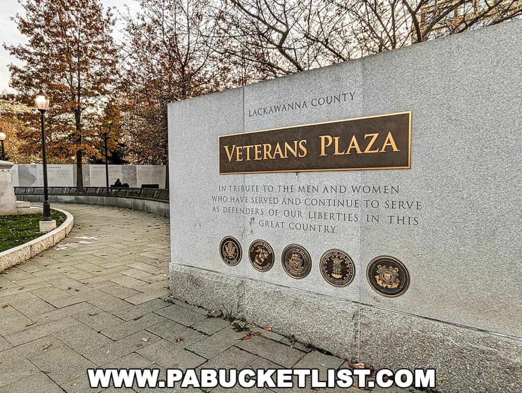 The Lackawanna County Veterans Plaza in Scranton, Pennsylvania, features a large stone wall with a bronze plaque reading 'VETERANS PLAZA' and an inscription paying tribute to the men and women who have served and continue to serve as defenders of liberties in the United States. Military service medallions are displayed below the inscription. A curved bench and leafless trees set against a dusk sky create a serene atmosphere.