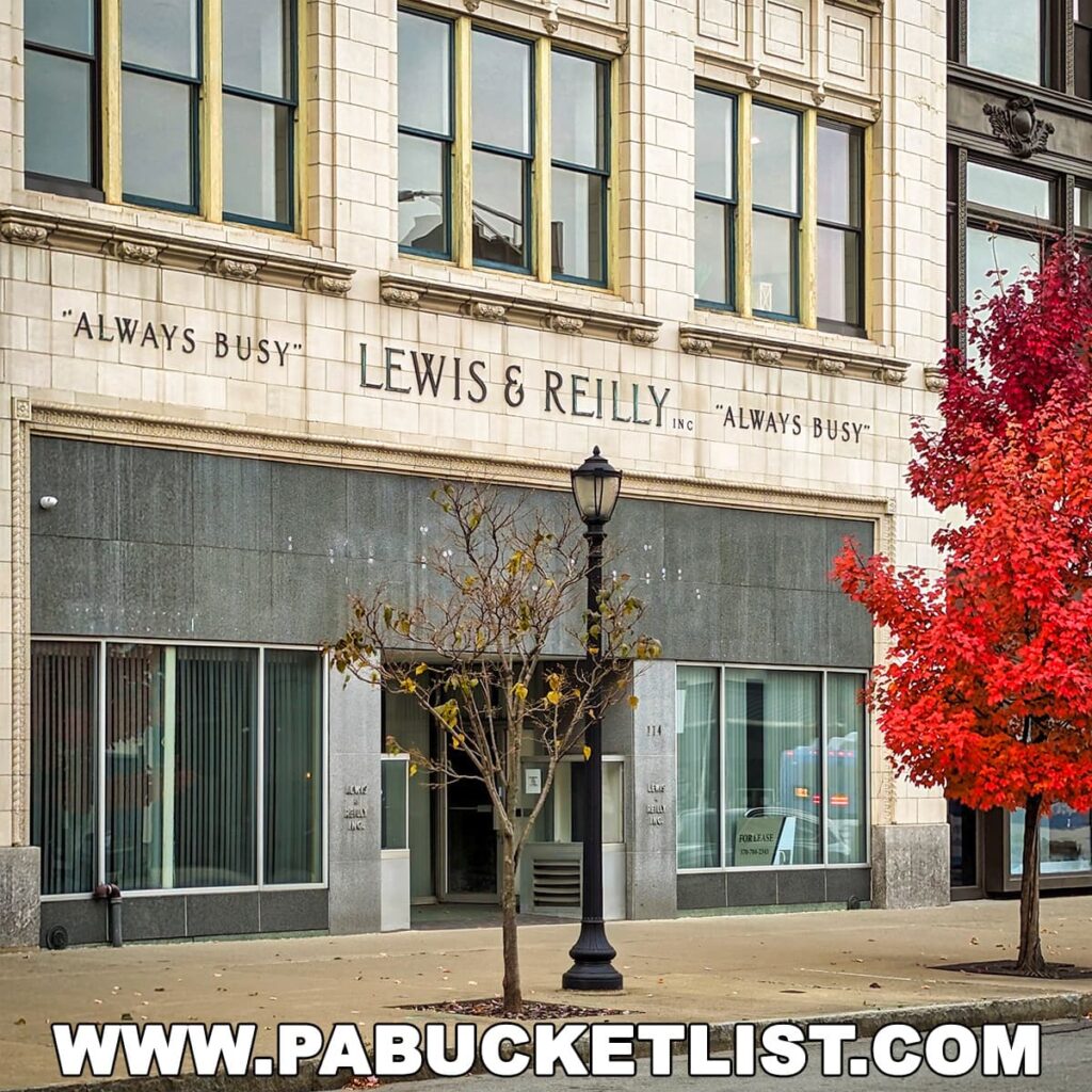 The façade of the 'Lewis & Reilly' building in downtown Scranton, Pennsylvania, with the phrase 'ALWAYS BUSY' inscribed above the large windows on its white terra cotta exterior. The ground level features dark grey panels, and the entrance is flanked by frosted glass windows. A street lamp and a young tree with sparse leaves stand in front, with a vibrant red tree to the right indicating autumn.