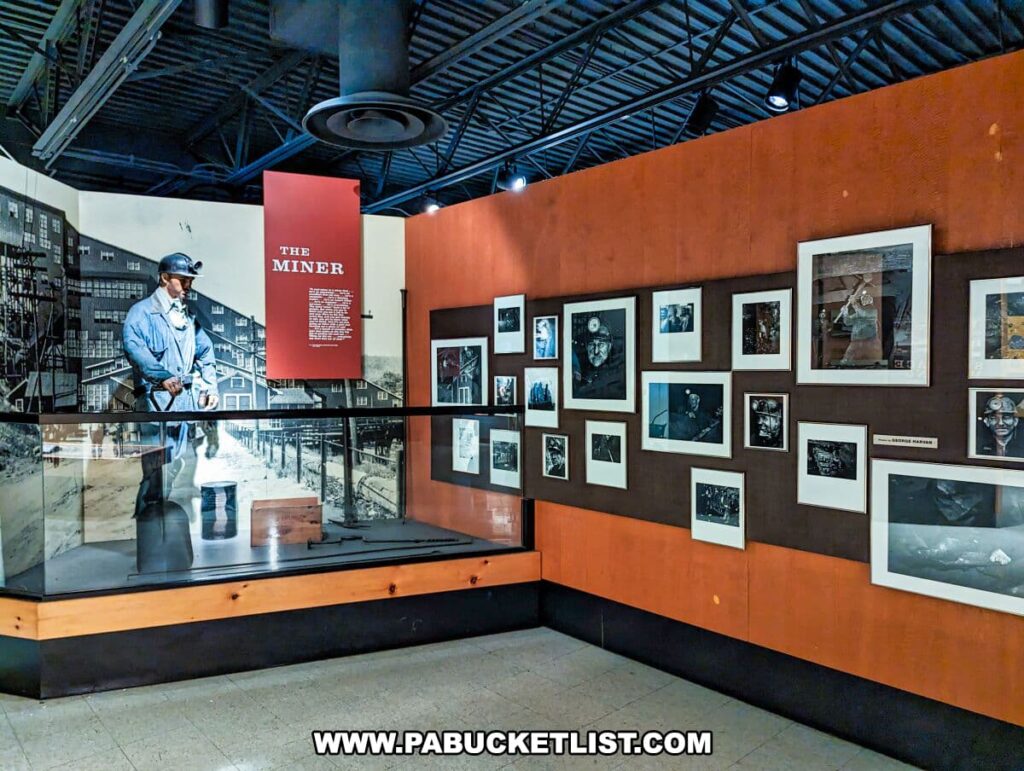 Inside the Museum of Anthracite Mining in Ashland, PA, an exhibit dedicated to 'THE MINER' is showcased. A life-sized mannequin of a miner wearing a hard hat with a headlamp, blue work clothes, and carrying a lunch pail stands in a glass case. Behind it, a large red panel with white text tells the story of the miner. The surrounding walls are lined with framed black and white photographs depicting various aspects of coal mining life and work. The industrial ceiling with exposed ductwork adds to the ambiance of the exhibit.