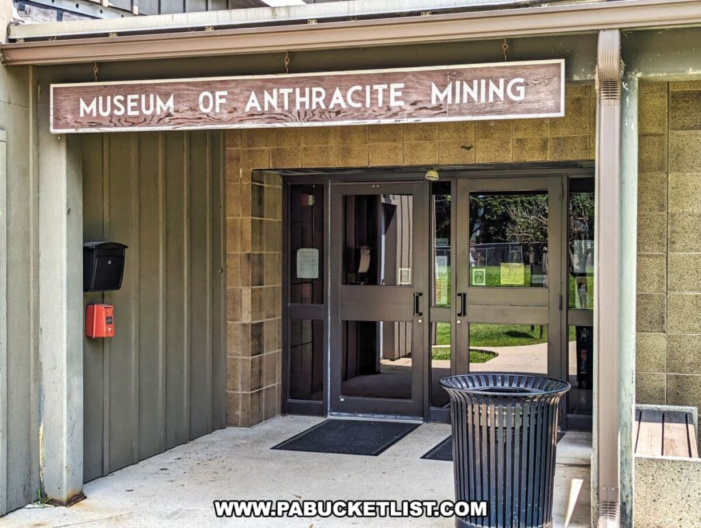 Close-up view of the entrance to the Museum of Anthracite Mining in Ashland, PA. Above the double glass doors, a weathered wooden sign with white letters states 'MUSEUM OF ANTHRACITE MINING.' To the left of the entrance, there is a black mailbox and a red fire alarm box mounted on the beige vertical siding.