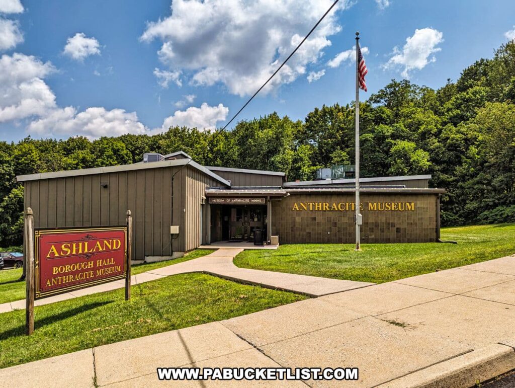 Exterior view of the Ashland Borough Hall Anthracite Museum, nestled in a lush green setting under a blue sky with scattered clouds. The museum building is a simple, one-story structure with a brown façade and a central entrance flanked by windows. In the foreground, a red and gold sign reads 'ASHLAND BOROUGH HALL ANTHRACITE MUSEUM.' An American flag on a flagpole stands proudly to the right of the building.