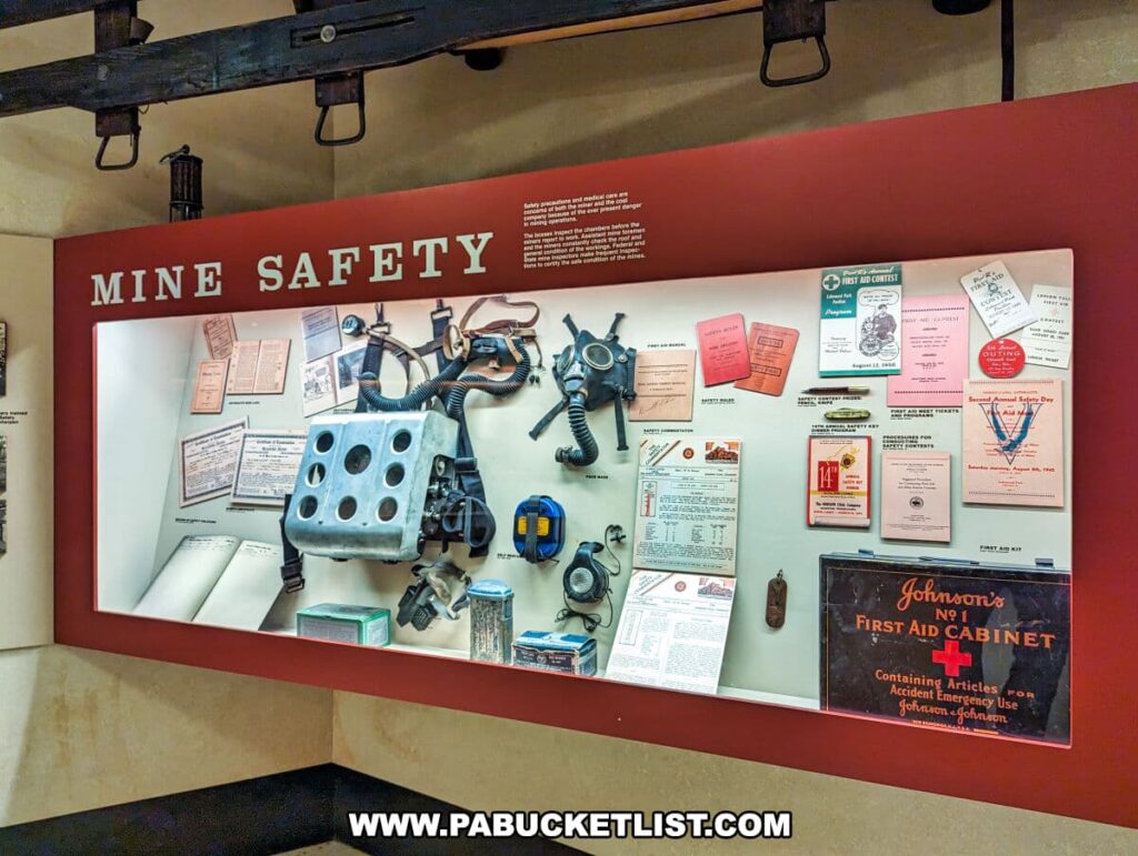 A 'MINE SAFETY' exhibit at the Museum of Anthracite Mining in Ashland, PA. The display, against a red background, features a variety of safety equipment and informational documents. Included are breathing apparatuses, a vintage 'Johnson's No. 1 First Aid Cabinet,' and several safety checklists and certificates. Above the exhibit hangs a beam with an old-fashioned mining lantern, adding to the authenticity of the setting. Various safety-related tags and pamphlets are pinned to the board, providing visitors with a historical perspective on mine safety measures.