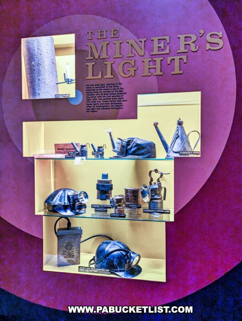 Exhibit titled 'THE MINER’S LIGHT' at the Museum of Anthracite Mining in Ashland, PA. Various types of historic miners' lights are displayed on yellow shelves against a purple wall. The collection includes a 'MINER’S SUNSHINE' lamp, different models of carbide lamps, and a safety hat with an attached electric cap lamp. Each piece is accompanied by a small descriptive tag. The lighting in the display case enhances the metallic surfaces of the lamps, reflecting the ingenuity and evolution of mining technology.