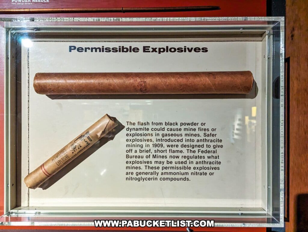 A display case at the Museum of Anthracite Mining in Ashland, PA, entitled 'Permissible Explosives.' It contains a cylindrical stick of dynamite and a wrapped cartridge labeled 'Permissible Explosive.' Below the items is an informative text explaining that safer explosives, introduced into anthracite mining in 1909, were designed to give off a brief, short flame to prevent mine fires or explosions in gaseous mines. It also mentions that the Federal Bureau of Mines regulates the use of explosives in anthracite mines, which generally contain ammonium nitrate or nitroglycerin compounds.