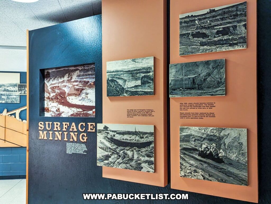 An educational wall display on 'SURFACE MINING' at the Museum of Anthracite Mining in Ashland, PA. The exhibit consists of several black and white historical photographs mounted on a blue and coral panel, each with descriptive texts that provide insights into the evolution of surface mining techniques. The images show large-scale operations with steam shovels and other machinery used in the past, and the captions explain the transition to diesel-powered equipment in modern times. The bold title 'SURFACE MINING' is featured prominently on the wall, guiding museum visitors through the history of coal extraction methods.