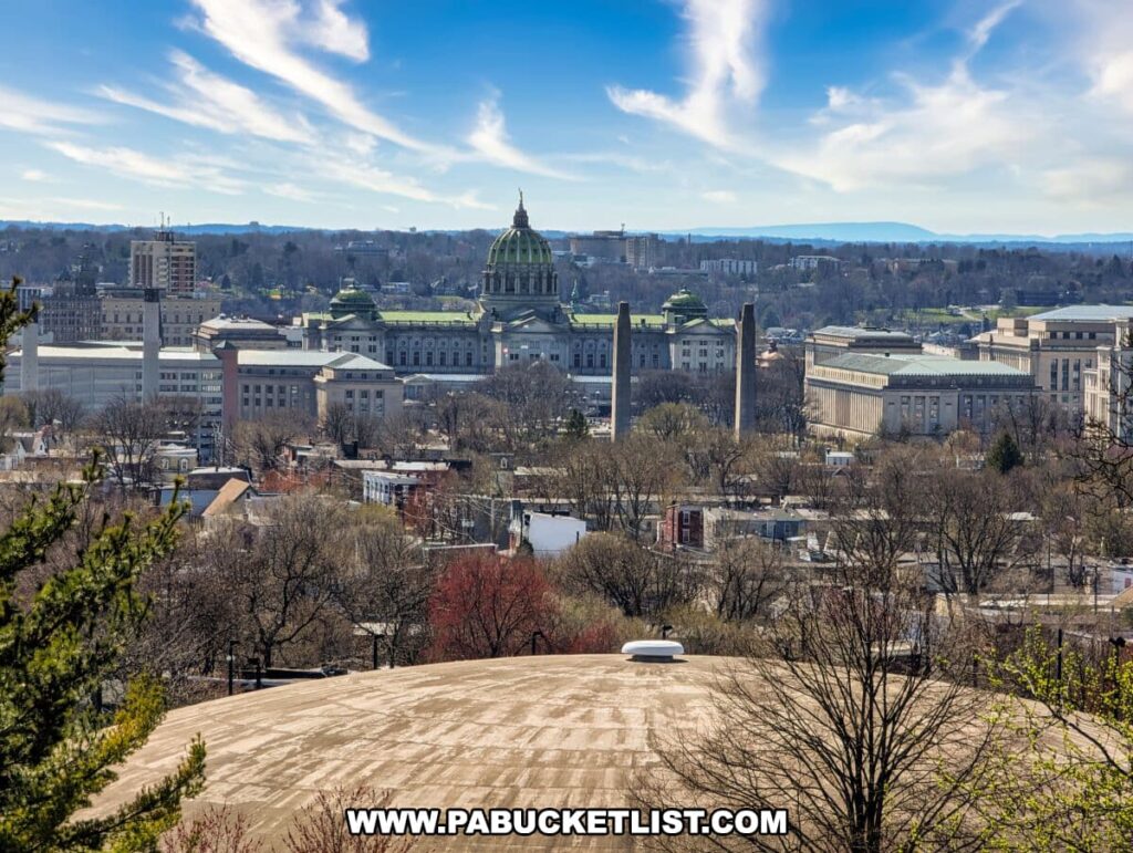 The image captures a scenic view from The National Civil War Museum in Harrisburg, PA, overlooking the Pennsylvania State Capitol building. The Capitol's green dome stands out amidst the surrounding cityscape, with various other government buildings and urban architecture in view. The landscape is dotted with trees, some of which are in the foreground, and a clear blue sky with a few clouds stretches above the serene setting. The vantage point offers a broad panorama of Harrisburg and highlights the grandeur of the Capitol in the context of the city.