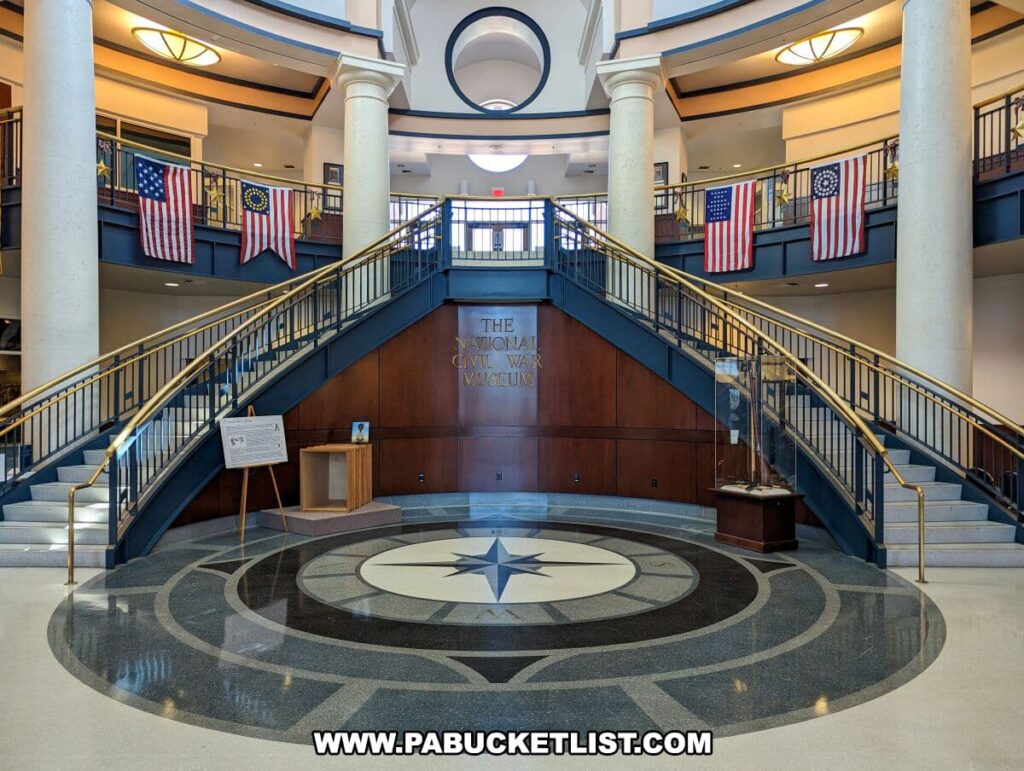 The image displays the grand foyer of The National Civil War Museum in Harrisburg, Pennsylvania. A sweeping dual staircase leads to a second-level balcony, where multiple American flags from different eras are hung. The foyer is spacious with a high ceiling, large columns, and a circular floor design featuring a star motif at the center. The words "The National Civil War Museum" are prominently displayed on the wall above the staircase, welcoming visitors to the museum. The environment is well-lit, with natural light streaming in, creating an inviting space for guests to begin their journey through the museum.