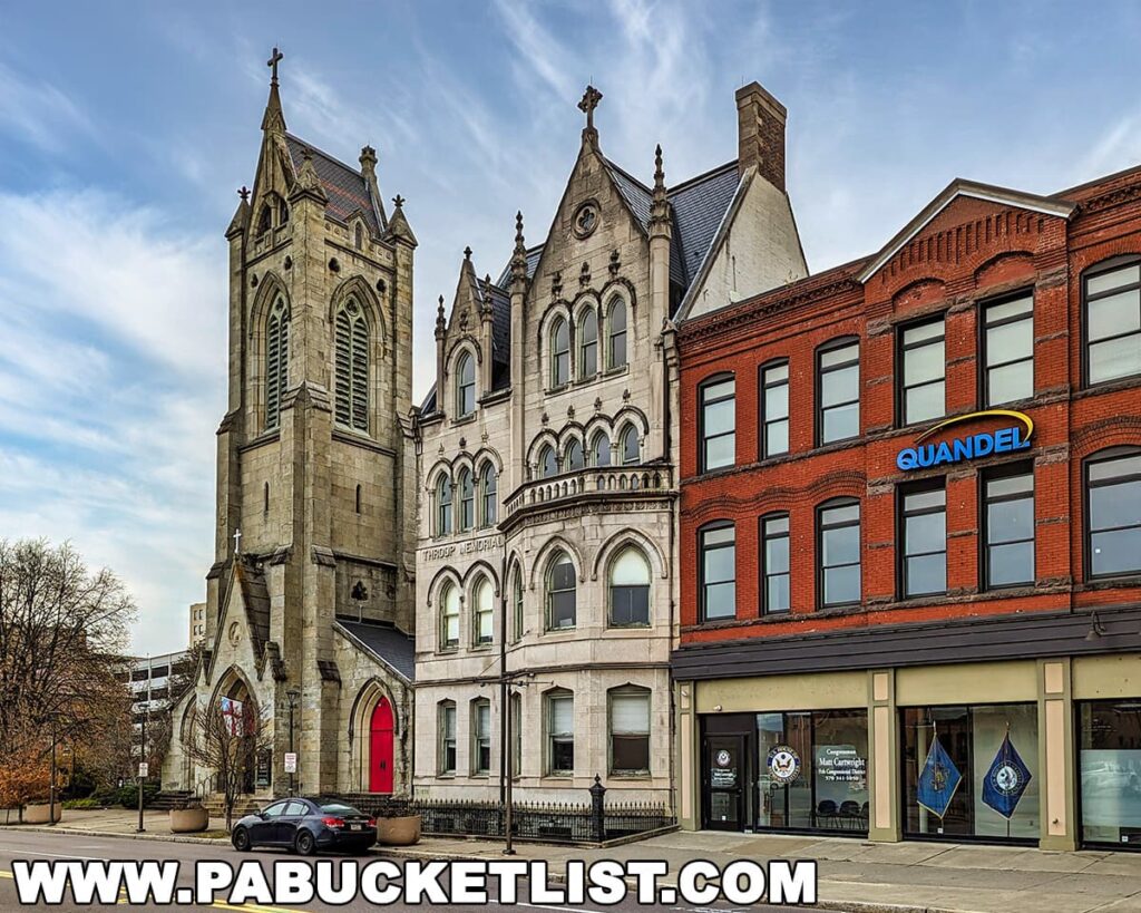 A view of the historic St. Luke’s Episcopal Church and its adjoining parish house in downtown Scranton, Pennsylvania, with their intricate Gothic Revival architecture, including pointed arches and a tall steeple. Next to the church, a row of red-brick commercial buildings with large windows, including one with a 'Quandel' sign, adds to the urban landscape.