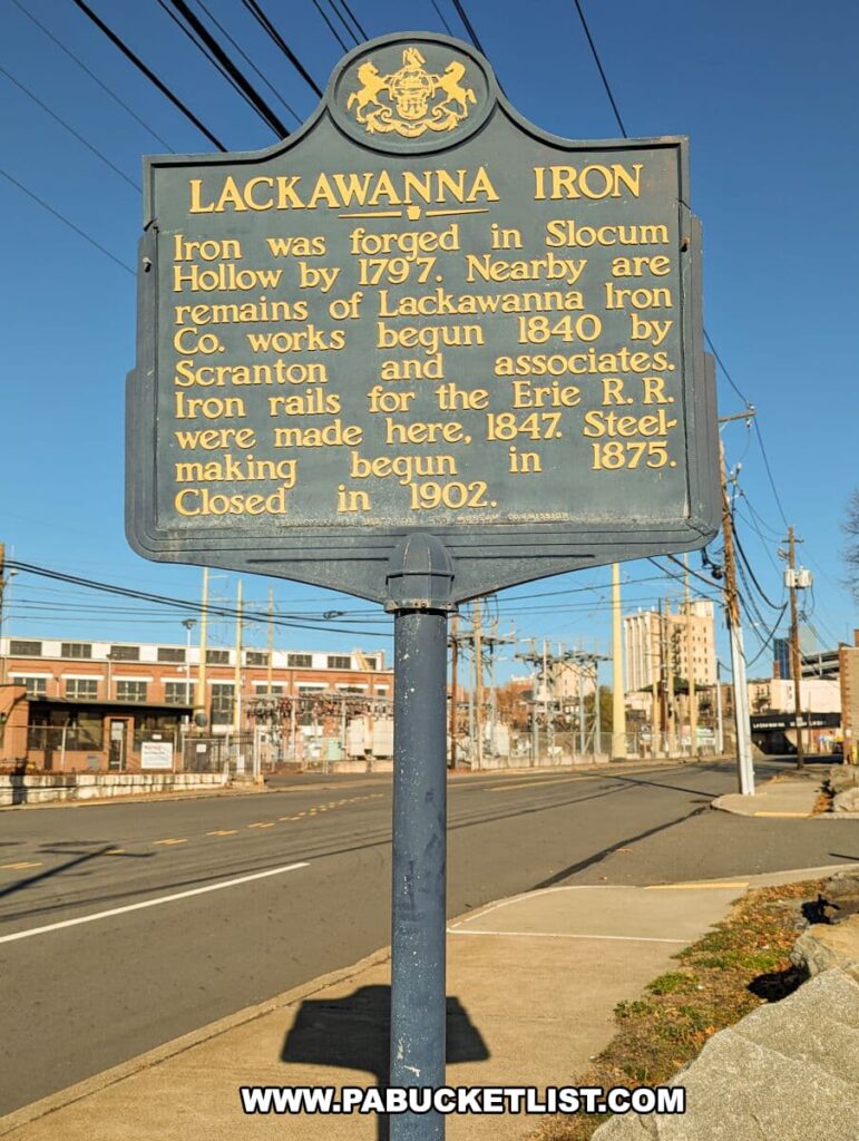 A historical marker for Lackawanna Iron in Scranton, PA, detailing the site's significance. The blue and yellow sign states that iron was forged in Slocum Hollow by 1797 and mentions the nearby remains of Lackawanna Iron Co. works begun in 1840. It notes that Scranton and associates made iron rails for the Erie Railroad here in 1847, and that steel-making began in 1875 and ended when the facility closed in 1902. The sign is set against a backdrop of a street with power lines and a building, captured on a sunny day with clear skies.