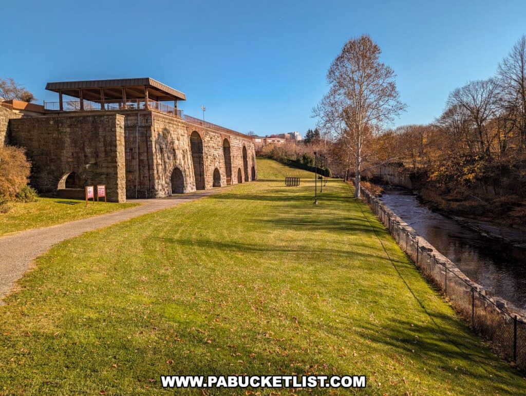 The Scranton Iron Furnaces Historic Site on a bright day, with a picturesque view of the stone structure and multiple arches. A modern, covered viewing platform sits atop the stone furnace remains, overlooking a lush green lawn. A paved path leads to the furnaces, and informational signs are visible at the path's entrance. To the right, Roaring Brook flows gently by, bordered by a chain-link fence, with leafless trees lining its banks against a backdrop of a clear blue sky.