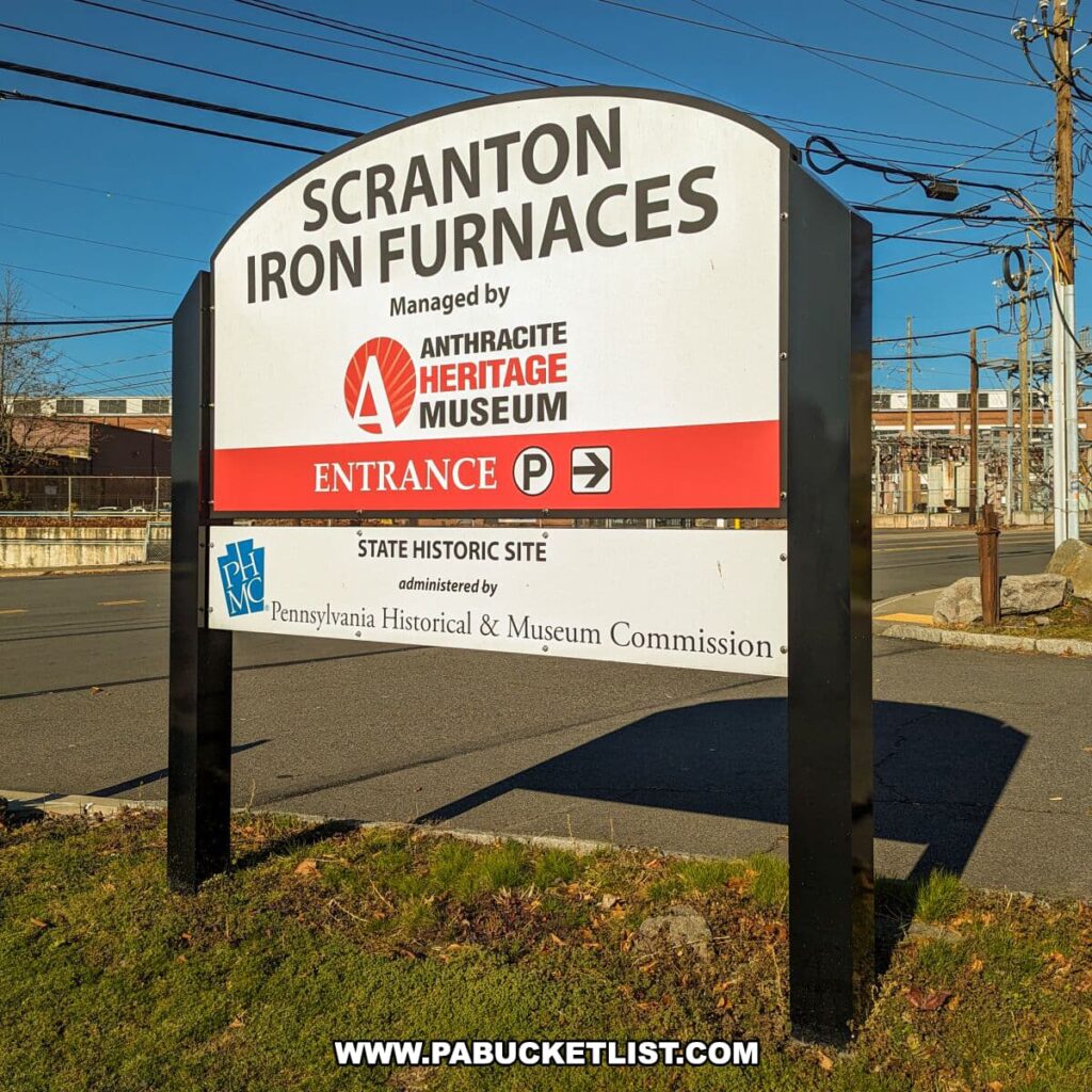Entrance sign for the Scranton Iron Furnaces in Scranton, PA, under a clear blue sky. The sign indicates the site is managed by the Anthracite Heritage Museum and is a state historic site administered by the Pennsylvania Historical & Museum Commission. It features symbols for parking and accessibility, with power lines in the background and a glimpse of industrial structures beyond.