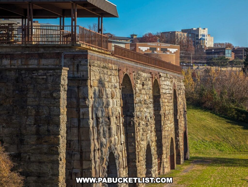 A side view of the Scranton Iron Furnaces Historic Site, with a large stone arch structure and a modern viewing platform on top. In the distance, the buildings of the University of Scranton rise above the treeline under a clear blue sky. The historic architecture of the furnaces contrasts with the modern campus buildings, illustrating the blend of historical and contemporary elements in Scranton, Pennsylvania.