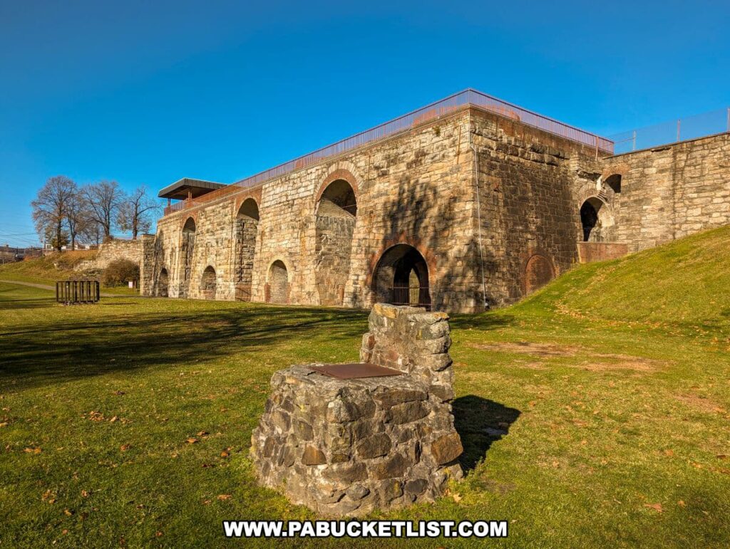 The historic Scranton Iron Furnaces in Scranton, PA, shown on a sunny day with clear blue skies. The image captures the stone structure of the furnaces, featuring multiple arched openings and a grassy hill in the foreground. A well-preserved stone well or monument is visible in the left foreground, and leafless trees dot the background. Shadows from the low sun stretch across the vibrant green lawn, highlighting the texture of the stone construction and the tranquility of the site.