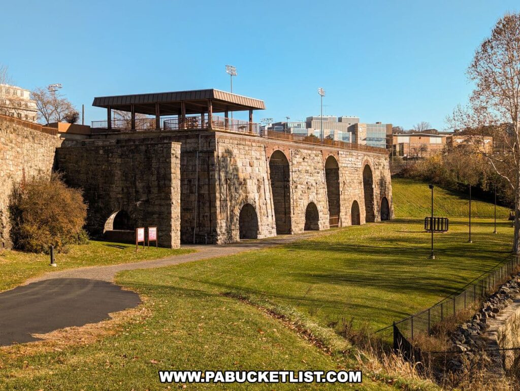 A sunny day view of the Scranton Iron Furnaces Historic Site in Scranton, PA, featuring a series of stone arches from the remaining furnace structures. A modern viewing platform with a roof is built atop the old stone foundation. In the foreground, a well-maintained grassy area and a walking path invite visitors, with informational signage visible to the left. Bare trees and a clear blue sky complete this tranquil historical scene.