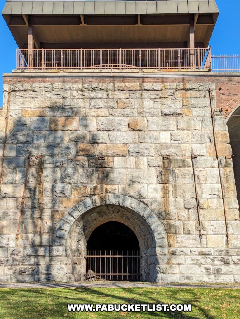 A ground-level view of one of the stone blast furnaces at the Scranton Iron Furnaces Historic Site, showing a large arched opening secured with a metal gate. Above the arch, the furnace's massive stonework supports a modern viewing platform with a metal railing. The clear blue sky in the background contrasts with the warm stone texture, and the green grass in the foreground adds to the peaceful setting.