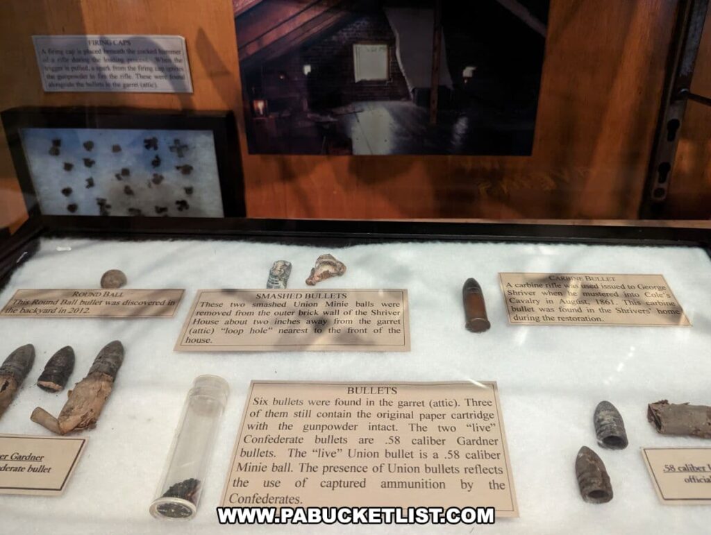 A glass display case at the Shriver House Museum in Gettysburg, Pennsylvania, contains a collection of Civil War relics. The exhibit includes a round ball bullet found in the backyard, smashed bullets from the brick wall of the Shriver House, and bullets recovered from the attic with their original paper cartridge with gunpowder intact. Also featured is a carbine bullet issued to George Shriver, who was mustered into Cole's Cavalry. Informative labels provide context for each item, explaining their significance and how they relate to the history of the house and the Battle of Gettysburg.