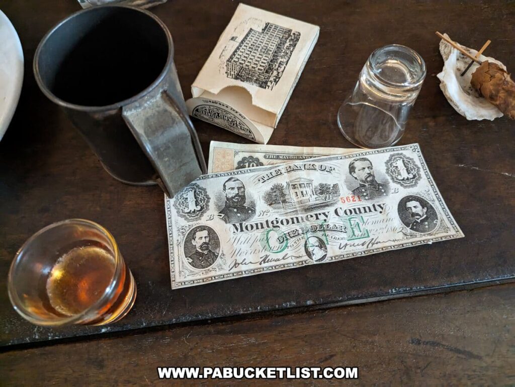 A close-up of a display on a wooden table at the Shriver House Museum in Gettysburg, Pennsylvania, showing items from the Civil War period. Central to the photo is a piece of historical currency, specifically a bill from "The Bank of Montgomery County," with portraits of men from the era and intricate detailing. Beside the bill is an empty glass salt shaker, a glass with a small amount of amber liquid, and a pewter mug. Behind these items is a period newspaper, adding to the authenticity of the scene. The display is arranged to give visitors a sense of the everyday items used during the time.