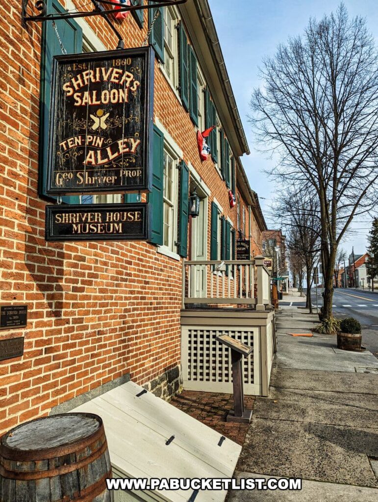 A historic brick building with green shutters, housing the Shriver House Museum in Gettysburg, Pennsylvania. A wooden sign for "Shriver's Saloon Ten-Pin Alley" hangs prominently, indicating the building's 1860 establishment. Below, a smaller sign confirms "Shriver House Museum." American flags adorn the second-story windows, and a barrel sits beside the entrance ramp.