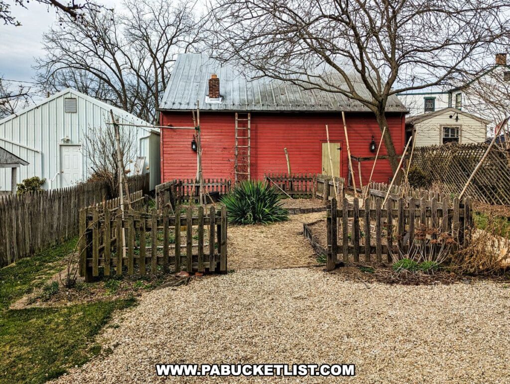 A rustic backyard garden at the Shriver House Museum in Gettysburg, Pennsylvania. The garden, enclosed by a weathered wooden picket fence, features early spring vegetation and bare branches, hinting at the onset of growth. A graveled pathway leads to a red outbuilding with a metal roof, contrasting with the white exterior of an adjacent structure. A tall, leafless tree stands guard over the space, and a series of tools and ladders suggest maintenance or historical farming practices. The setting provides a glimpse into the domestic life of the era represented by the museum.