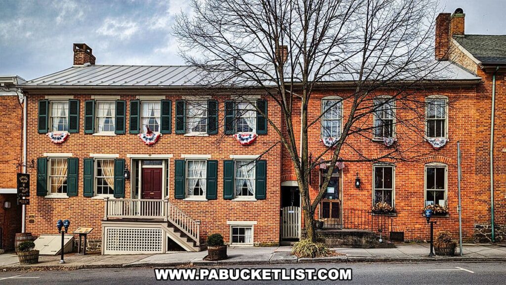 The front facade of the Shriver House Museum in Gettysburg, Pennsylvania, is captured in this image. It features a two-story red brick building with green shutters and a metal roof. Each window on the second floor is adorned with patriotic bunting, and a leafless tree stands in front, suggesting the photo was taken in a cooler season. The entrance has a red door accessible by a set of steps with a ramp, and a small sign beside the door indicates the building's status as a museum. A quiet street setting gives the historic structure a quaint and inviting appearance.