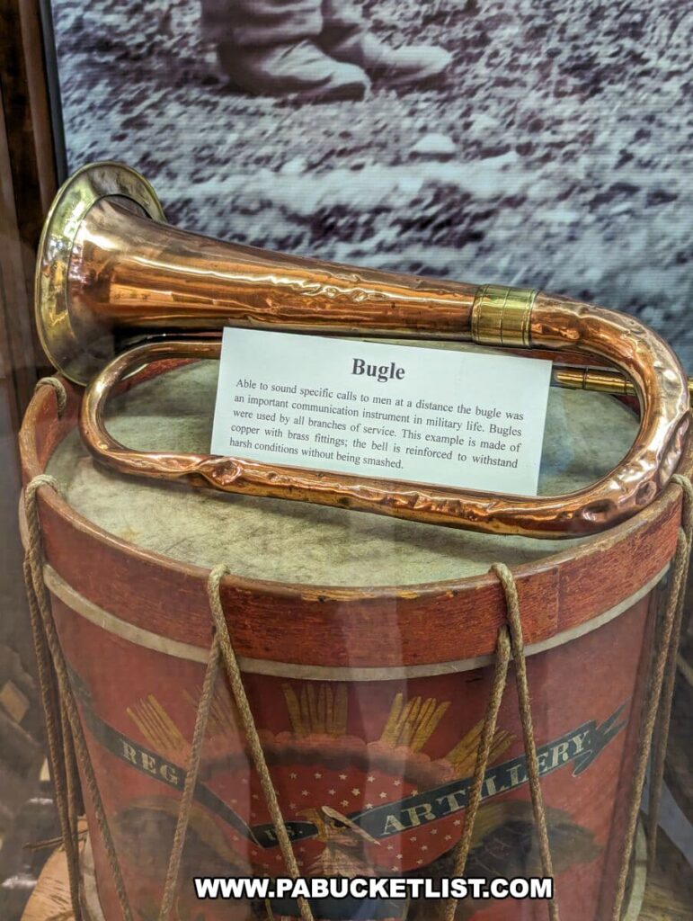 Historic brass bugle displayed on top of a red and gold decorative military drum at the Soldiers and Sailors Memorial Hall and Museum in Pittsburgh, PA. The bugle, used to signal calls to soldiers, rests against a background of a faded black and white photograph. A label in front of the bugle explains its significance as an important communication instrument in military life, mentioning its reinforcement to withstand harsh conditions.
